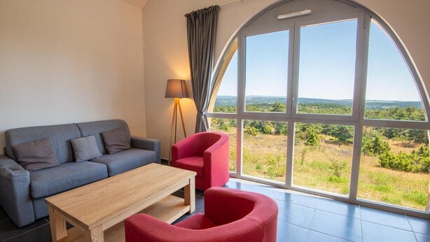 A modern living room with a grey sofa, a wooden coffee table, and red chairs. A floor lamp and a large arched window provide natural light and a view of the outdoors. - La Baumelle 4/6 persons (SAINT-PIERRE-DES-TRIPIERS)