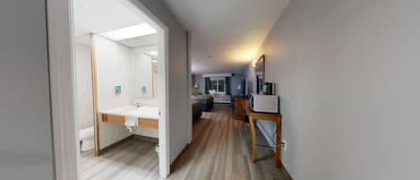 A hotel room with a wooden floor, a bed, a desk, a chair, a TV, a microwave, and a bathroom with a sink and mirror.