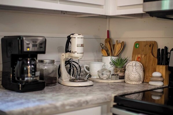 Una encimera de cocina con una cafetera, un robot de cocina, un tarro, una planta pequeña, un salero, una tabla de cortar de madera y un bloque para cuchillos.