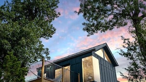 Eine moderne Holzhütte mit einer Holzterrasse und einem großen Fenster, umgeben von Bäumen und felsigem Boden.