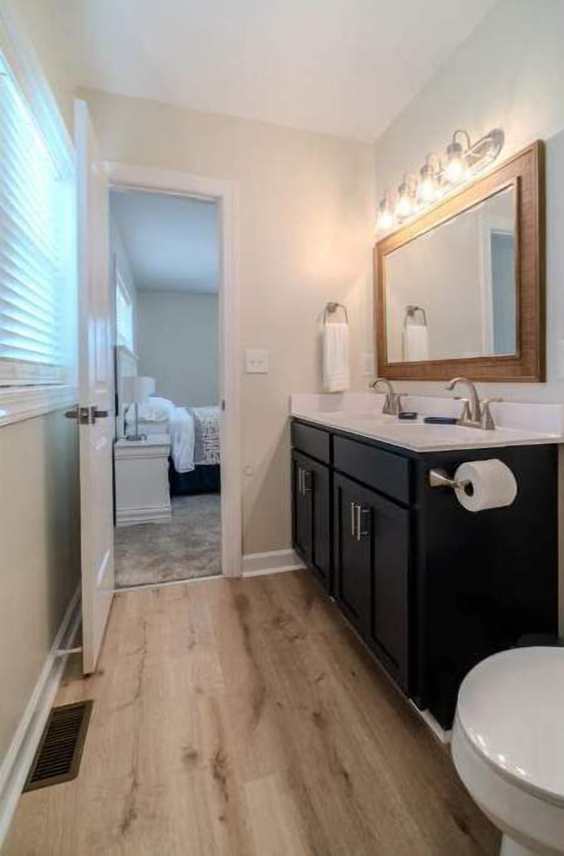 Une salle de bain avec un plancher en bois, des toilettes blanches, un meuble-lavabo noir avec un miroir et un espace douche visible à travers une porte ouverte.