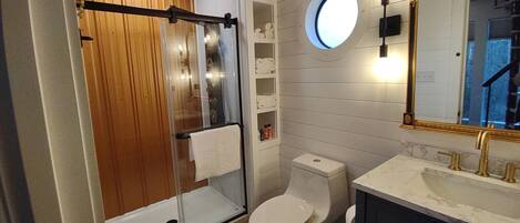 Un baño con cabina de ducha de vidrio, inodoro, lavamanos con detalles dorados y espejo redondo con marco dorado.
