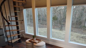 A room with a wooden staircase, a cushioned bench, ottomans, and a large window with a view of a wooded area. - The Nest, unplug& reconnect with that special someone in our charming birdhouse! (Troy)