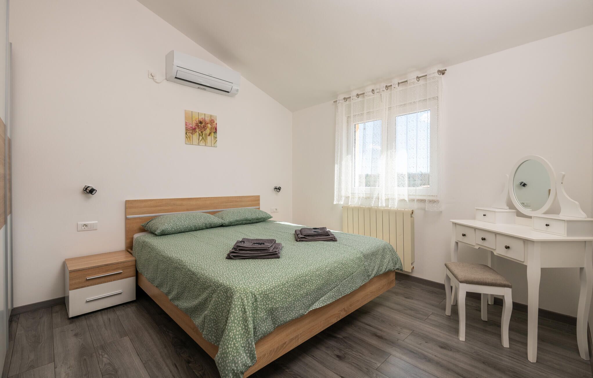 Un dormitorio con cama, escritorio con espejo, silla, aire acondicionado y ventana con cortinas.