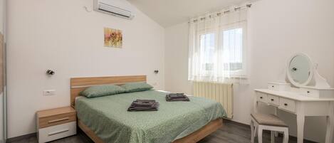 Un dormitorio con cama, escritorio con espejo, silla, aire acondicionado y ventana con cortinas.