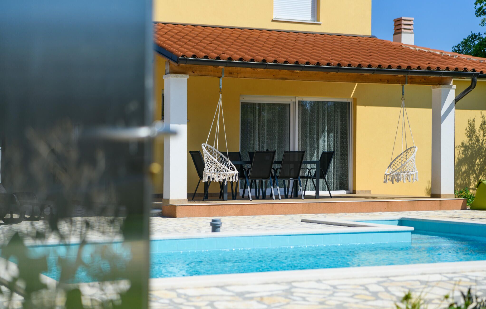 A poolside area with a covered seating area, a pool, and a yellow building with a tiled roof.