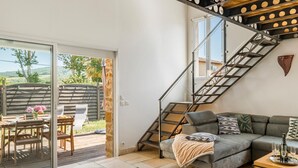 A modern living room with a grey sofa, a wooden dining table with chairs, a staircase with a metal railing, and a view of the outdoors through large glass doors.