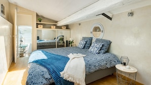 A bedroom with a skylight, a bed with patterned bedding, a bedside table with a lamp, and a round mirror on the wall.