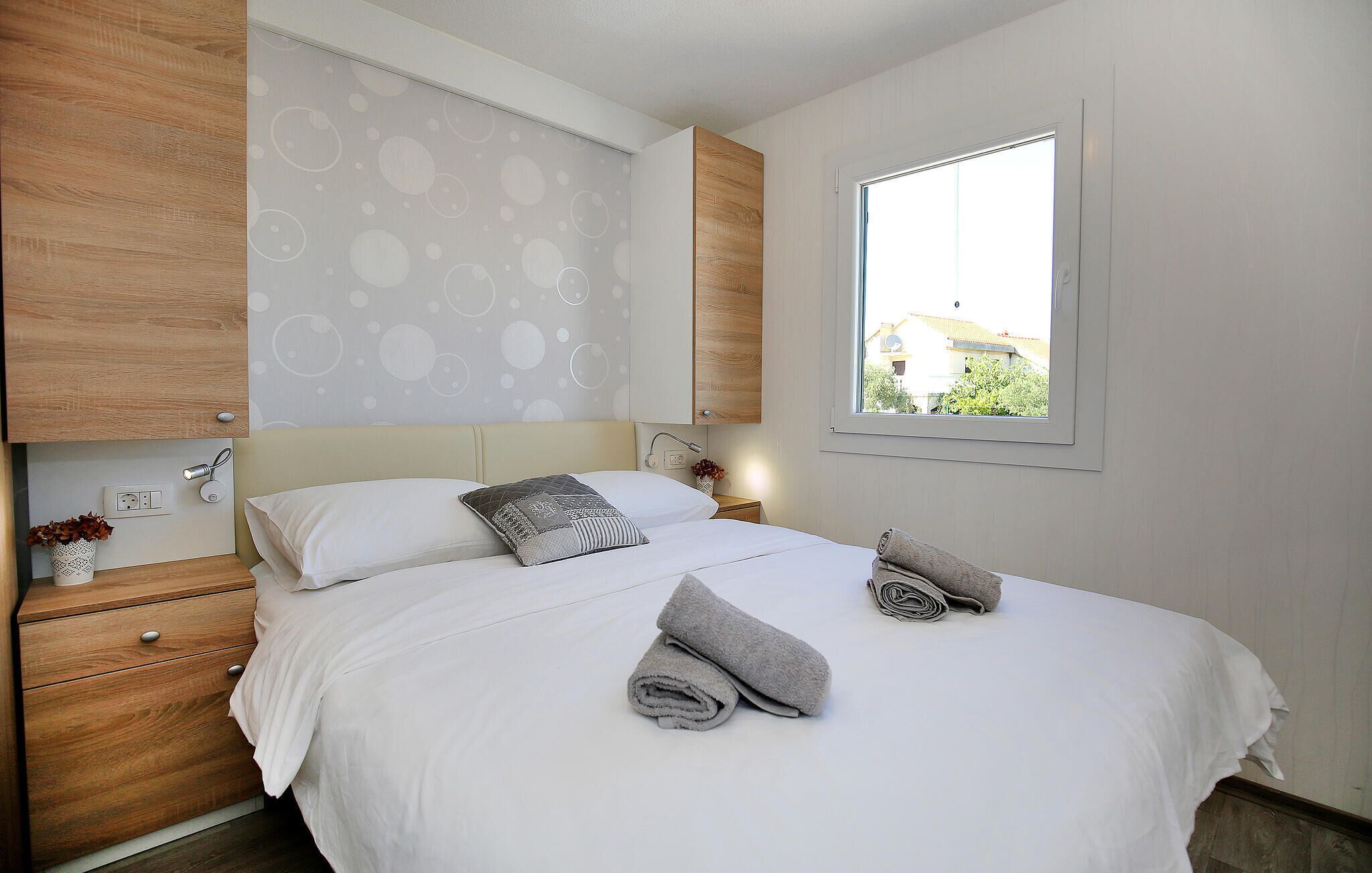 A neatly made bed with white linens and gray towels, a wooden nightstand with a plant, and a window with a view of buildings.