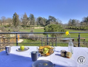 Ein gedeckter Tisch mit Obstkorb, Tasse, Glas und Flasche, mit Blick auf eine reizvolle Landschaft mit Bäumen und einem Gebäude im Hintergrund.