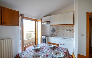 Een kleine keuken met witte kastjes, een kookplaat en een spoelbak. Er staat een eettafel met een tafelkleed met bloemen en borden.