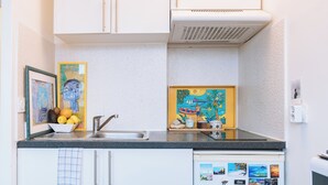 A kitchen with white cabinets, a black countertop, and a sink. There are two framed artworks on the wall, one depicting a blue figure and the other a colorful scene with a boat. - Stylish Studio 12 Min from Republique (Paris)