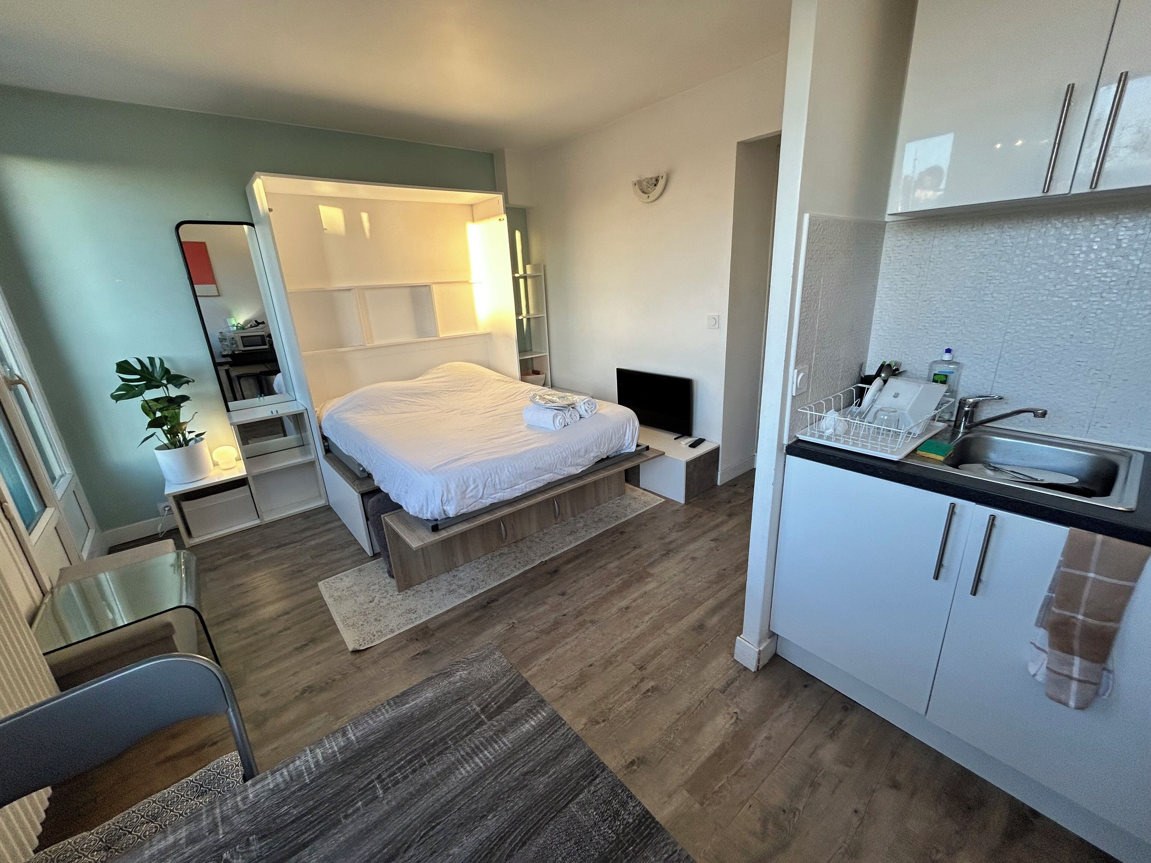 Una habitación de hotel moderna con cama, mesita de noche, televisor y una kitchenette con lavamanos y armarios.