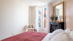 Ein Schlafzimmer mit einem großen Bett, einem Sessel, einem Schminktisch und einem Fenster mit Blick auf Gebäude.