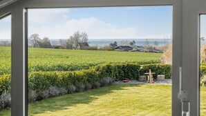 Udsigt gennem et vindue af et græsstykke med en trælegeplads, en hæk og en række huse ved havet.