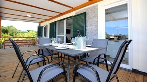 An outdoor dining area with a table set for four, surrounded by chairs, on a wooden deck with a stone wall and green shutters in the background.