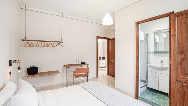 Un dormitorio con cama, escritorio, silla, lámpara colgante y baño visible a través de una puerta abierta.