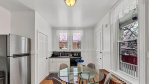 A compact kitchen with white cabinetry, a stainless steel refrigerator, a glass dining table with chairs, and a view of a snowy landscape through a window. - The Victorian on South Park • Sleeps 6 (Buffalo)