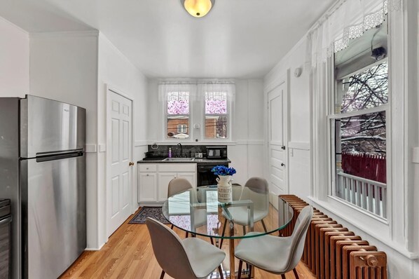 A compact kitchen with white cabinetry, a stainless steel refrigerator, a glass dining table with chairs, and a view of a snowy landscape through a window. - The Victorian on South Park • Sleeps 6 (Buffalo)