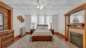 A bedroom with a large bed, a wooden dresser, a fireplace, and a ceiling fan. - The Victorian on South Park • Sleeps 6 (Buffalo)