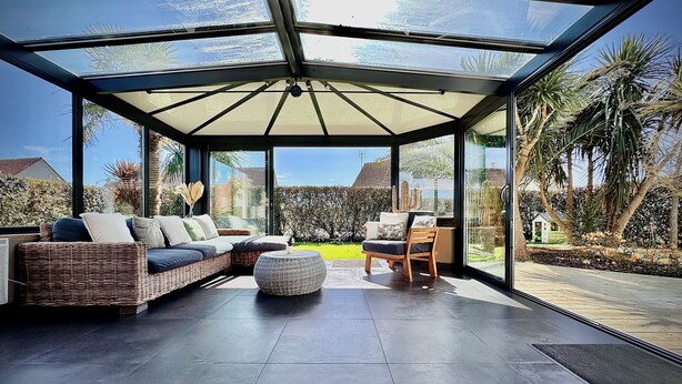 Ein moderner, abgeschlossener Patio-Bereich mit Glasdach, Rattan-Sofa, Holzstuhl und rundem Ottoman.