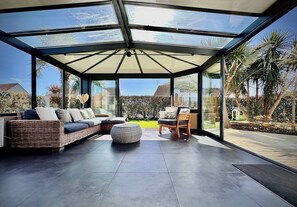 Un espace patio moderne et clos, doté d'un plafond en verre, d'un canapé en osier, d'une chaise en bois et d'un pouf rond.