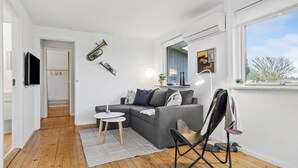 A modern living room with a grey sofa, a small round table, and a black chair. There is a flat-screen TV mounted on the wall, a window with a view of trees, and a door leading to another room. - 3 bedroom amazing home in Otterup (Otterup)