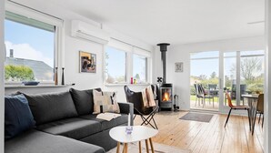 A modern living room with a grey sofa, a white coffee table, and a black chair. There is a fireplace and a large window with a view of the outdoors. - 3 bedroom amazing home in Otterup (Otterup)