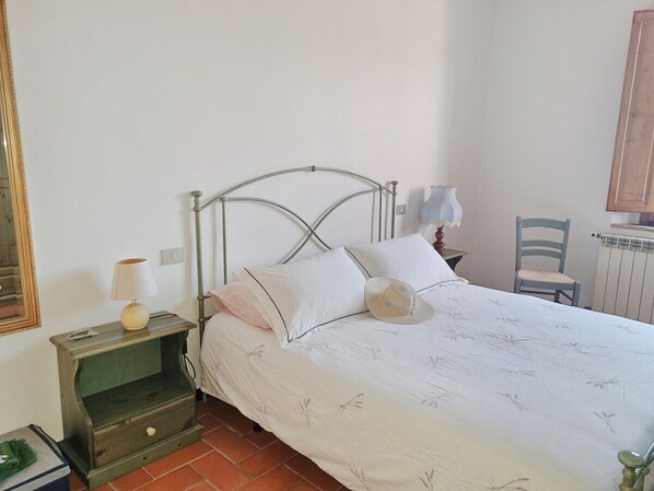 Un dormitorio con una cama de estructura metálica, una mesita de noche con lámpara, una silla y un radiador.