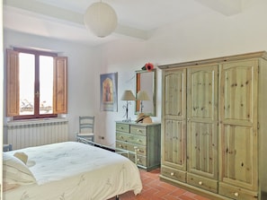 A bedroom with a bed, a dresser, a chair, a window with shutters, and a wardrobe.