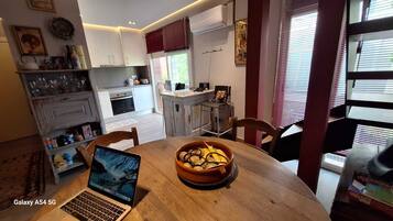 Una cocina compacta con armarios blancos, una mesa de comedor con una computadora portátil y un tazón de fruta, y una ventana con persianas.