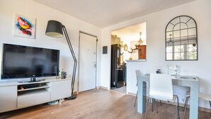 A modern living room with a flat-screen TV, a standing lamp, and a dining area with a glass table and chairs. - Bel appart avec balcon et clim près des Calanques (Marseille)