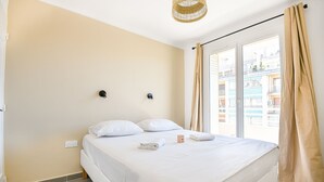 Un dormitorio con cama, dos lámparas de pared, un ventanal con cortinas y vistas a edificios.