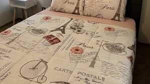 Ein Bett mit Paris-Motiv auf der Bettdecke, ein kleiner Nachttisch mit Lampe und ein gerahmtes Bild an der Wand.