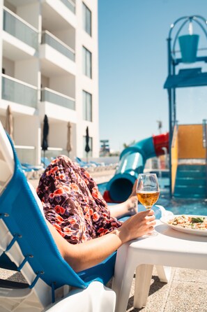 A person relaxing by a poolside with a glass of drink and a plate of food, a blue chair, and a building in the background. - High Garden Grand Hotel (Mazatlán)