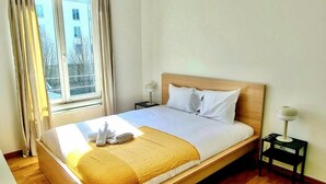 A neatly made bed with a wooden headboard, a bedside table with a lamp, and a window with curtains. - Luxury 1 bedroom flat w parking&balcony (Luxembourg City)