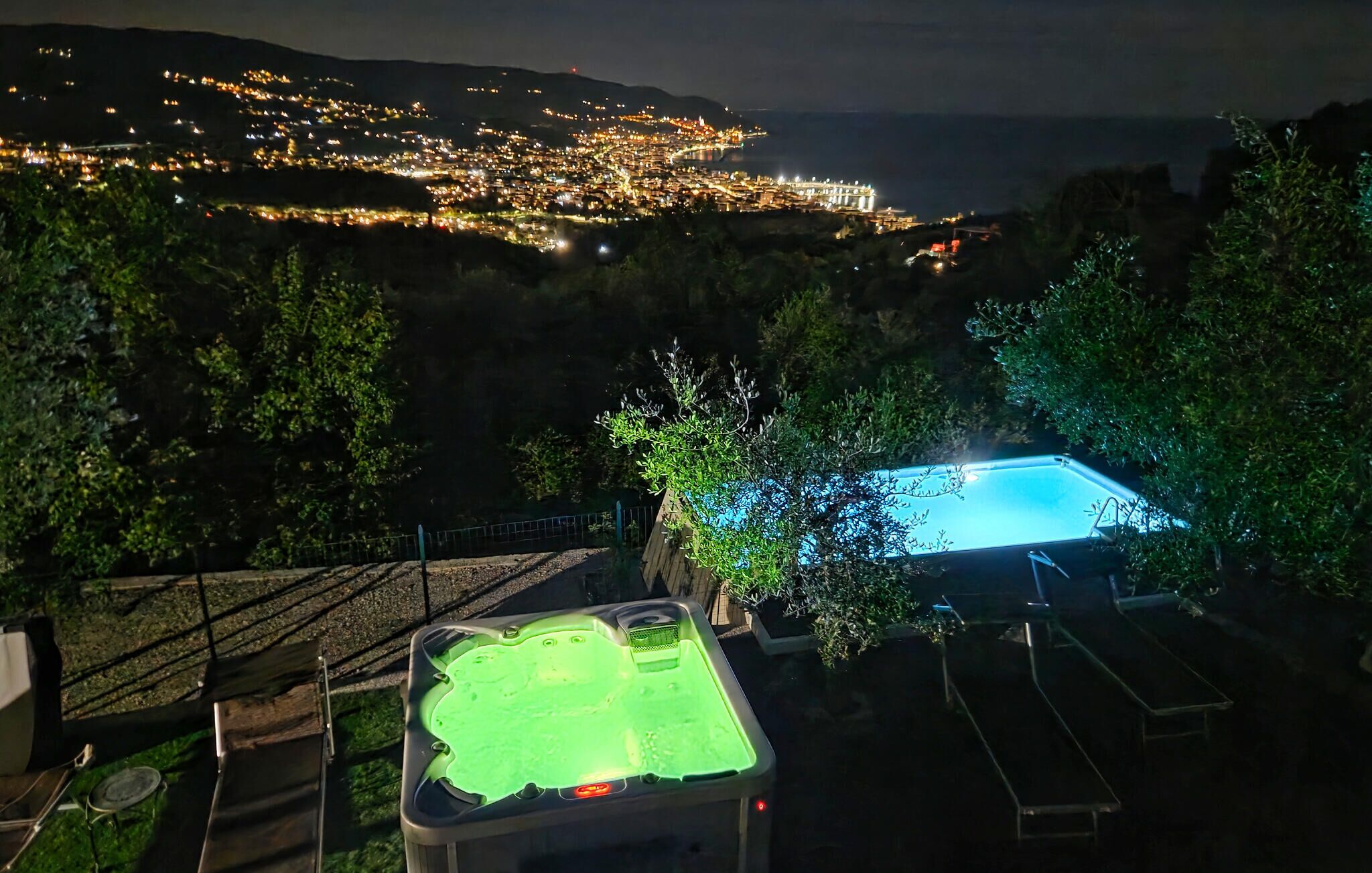 A pool area with a hot tub, surrounded by trees and a view of a cityscape at night.