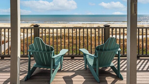 Een houten terras met twee groene Adirondack-stoelen, gericht op een strand met uitzicht op de oceaan.