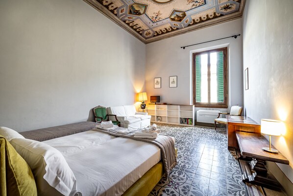 A hotel room with a bed, a desk, a chair, a lamp, and a window with shutters.