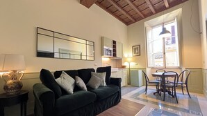 Uma sala de estar com um sofá verde escuro, uma mesa de centro de madeira, uma pequena mesa lateral com uma luminária, um espelho grande, uma mesa de jantar com cadeiras e uma janela com vista para prédios.