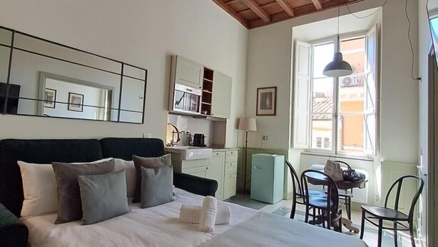 Quarto com cama grande, escrivaninha pequena, cadeira, janela e cozinha compacta.