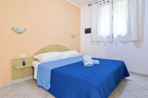 A bedroom with a bed, a nightstand, a wall-mounted light, and a window with curtains. - Two-room apartment of approximately 40 m2 composed of a living room with kitchenette, double sofa bed, double bedroom, bathroom, patio.Some apartments may also have a bunk bed.Endowed of:- TV- Outdoor chairs, table- External parking space- Kitchenette- A (Badesi)