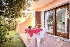 Un espace de restauration en plein air avec une nappe rouge, des chaises blanches et une porte vitrée donnant sur un jardin.