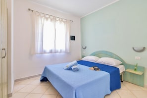 A single bed with a blue bedspread and white pillows, a small bedside table with a lamp, and a window with white curtains.