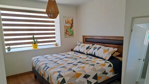 Une chambre à coucher avec un lit, une fenêtre avec des stores, un luminaire suspendu, une petite plante et une porte.