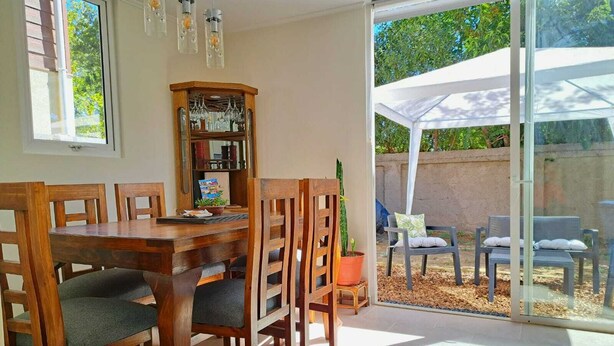 Un coin repas avec une table et des chaises en bois, une bibliothèque et une porte-fenêtre coulissante donnant sur une terrasse extérieure équipée de chaises et d’un auvent blanc.