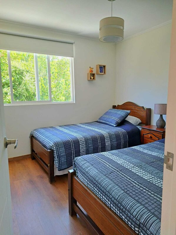 Un dormitorio con dos camas, una ventana que ofrece vistas a zonas verdes, una mesita de noche pequeña con una lámpara, y una estantería en la pared con objetos decorativos.