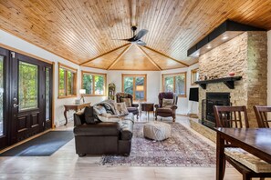 A spacious living room with a wooden ceiling, stone fireplace, leather sofa, and large windows. - Gimli Lakefront Cottage (Gimli)