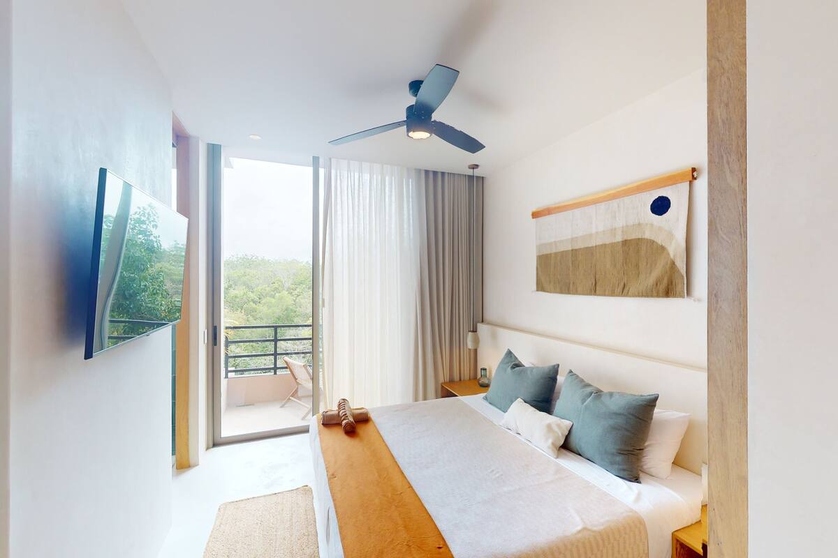 Quarto de hotel moderno com cama, escrivaninha, cadeira, ventoinha no teto e vista para o exterior.