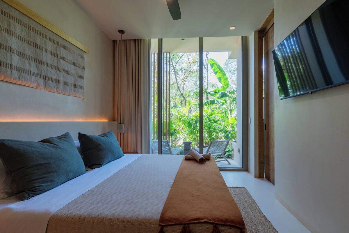 Quarto de hotel moderno com uma cama grande, televisão de tela plana e vista para vegetação exuberante através de janelas amplas.
